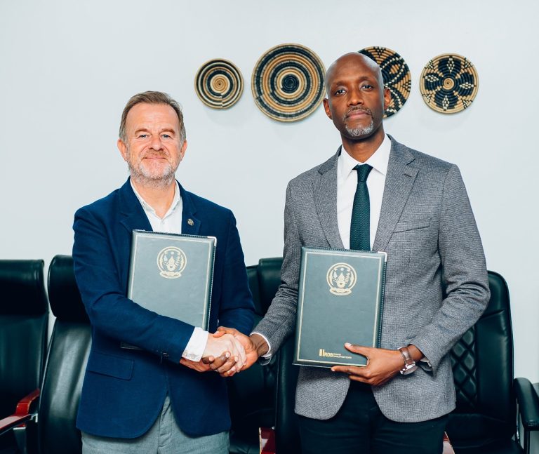 Two men in suits shake hands while holding official-looking folders in a meeting room with woven wall decor behind them.