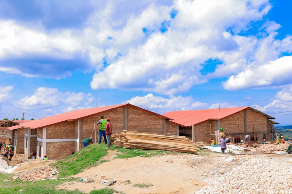 Construction site with brick buildings and red metal roofs; workers handling lumber and scaffolding on a dirt mound under a blue sky with clouds.