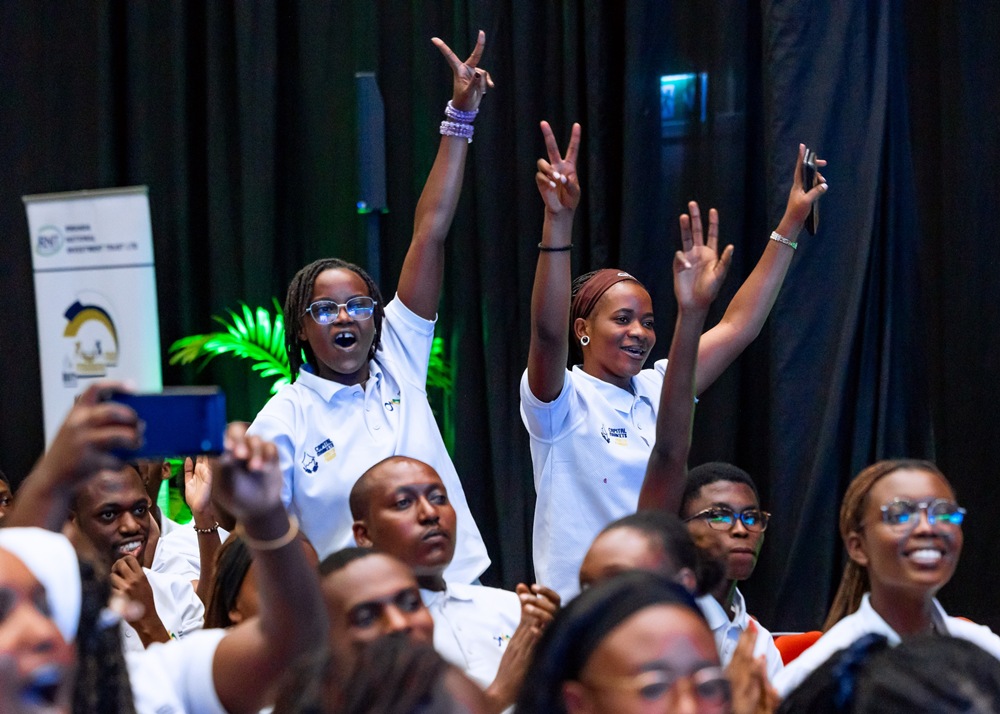 Group of diverse young adults at an indoor event, many raising their hands in celebration while seated in a crowd wearing white polo shirts.