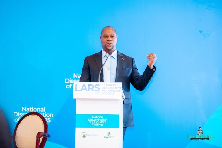 Man in a dark suit delivering a speech at a white podium against a bright blue backdrop, microphone in front, one arm raised