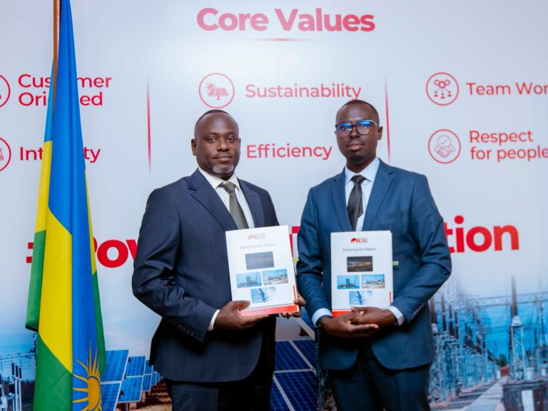 Two men in suits pose with brochures at a corporate event, backdrop displays Core Values and Sustainability icons and text.