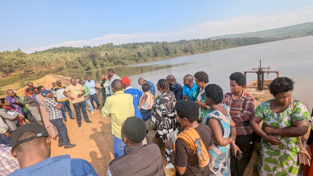 Group of people standing in a semicircle on a dirt riverside, talking together during a community gathering by the lake.