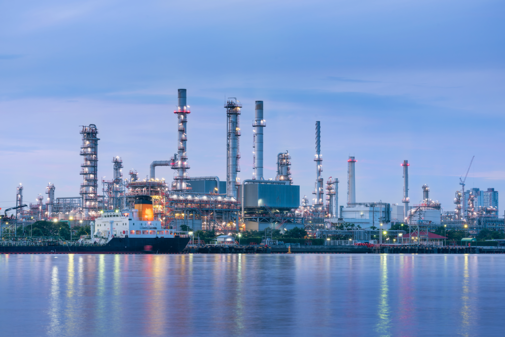 Waterfront oil refinery with tall processing towers lit at dusk, a docked ship visible in the foreground and reflections on calm water.