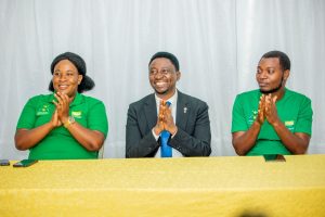 Three people sit at a yellow-clothed table, clapping and smiling during a formal event; two in green shirts flank a man in a suit in the center.