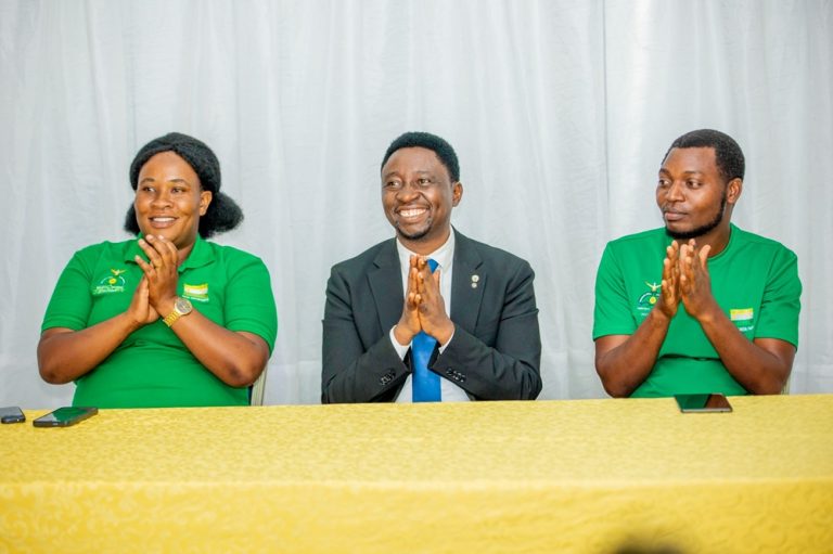 Three people sit at a yellow-clothed table, clapping and smiling during a formal event; two in green shirts flank a man in a suit in the center.