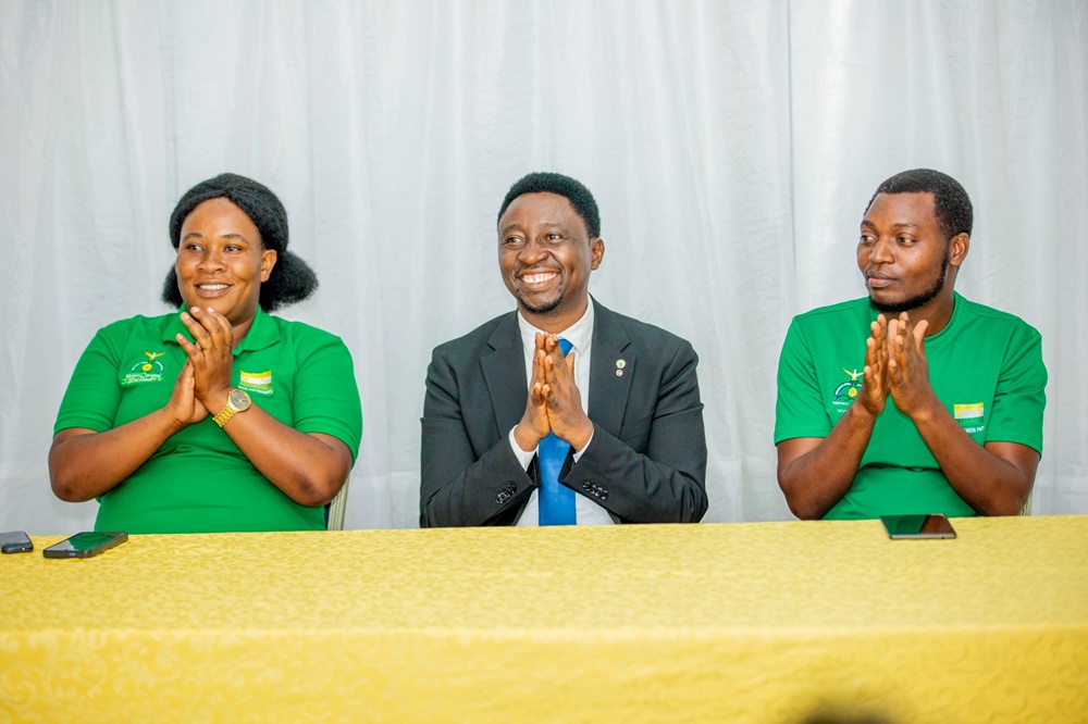 Three people sit at a yellow-clothed table, clapping and smiling during a formal event; two in green shirts flank a man in a suit in the center.