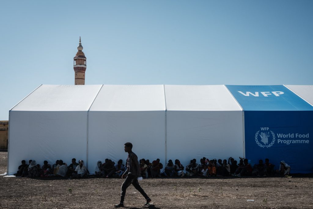 People seated under a large white WFP tent; a man walks by in the foreground, with a minaret and a clear blue sky in the background.