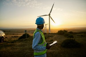 Worker in a blue hard hat and high-visibility vest checks a tablet near wind turbines at sunset in a desert landscape.