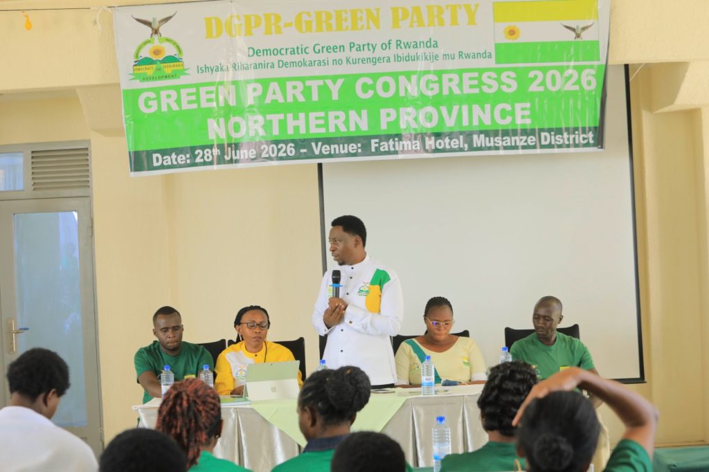 Man in white shirt with microphone addressing a panel at a Green Party congress banner backdrop