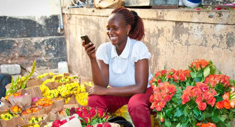 Smiling woman sits among colorful flower bouquets at an outdoor market, looking at her phone.