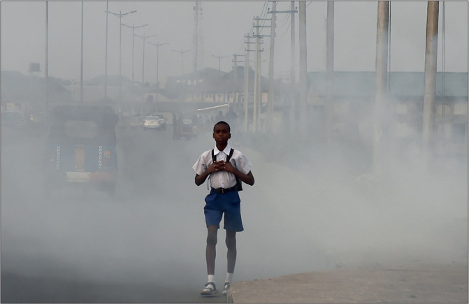 Boy in a school uniform walks along a smoke-filled street with vehicles fading in the hazy background.