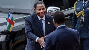 Two men in suits shake hands beside a black SUV; Comoros flag on the car and a uniformed guard nearby.
