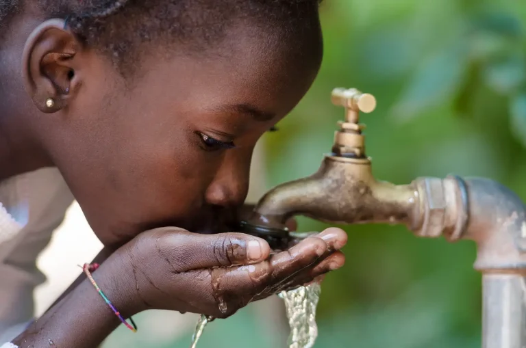 Young girl cupping water from an outdoor faucet, drinking directly from the tap amid a green blurred background.
