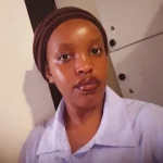 Close-up portrait of a woman wearing a brown head wrap and light blue collared shirt, looking at the camera.