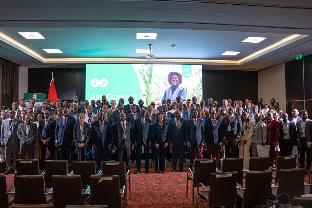 Group of diverse professionals in suits and dresses posing for a conference photo in a large meeting room, with an AgriConnect banner on the stage screen behind them.