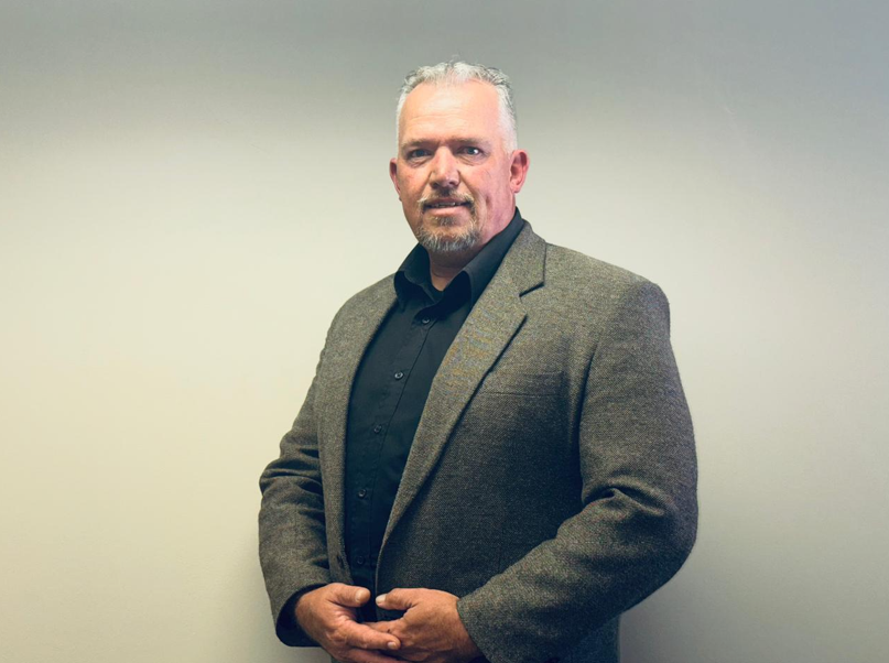 Professional man in a gray blazer and black shirt posing against a pale backdrop.