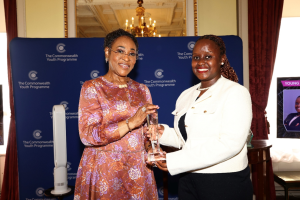 Two women smile as one awards a crystal trophy at a Commonwealth Youth Programme event, with a blue backdrop behind them.”]},{
