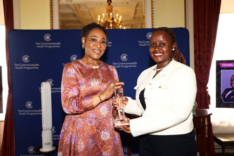 Two women smile as one awards a crystal trophy at a Commonwealth Youth Programme event, with a blue backdrop behind them.”]},{