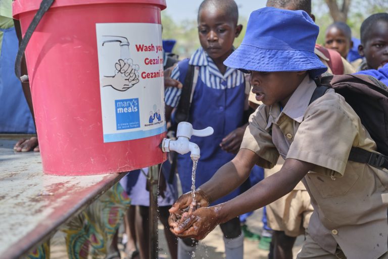 Children wash their hands at an outdoor water station with a pink bucket and running water, as onlookers stand nearby