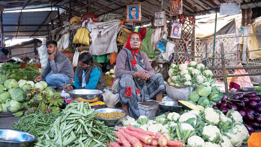 Central woman vendor in red headscarf sits amid fresh vegetables at a busy outdoor market stall.