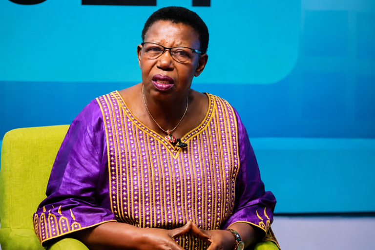 Woman wearing a purple and gold traditional dress sits on a lime-green chair, speaking on a panel with a lapel mic attached to her outfit.
