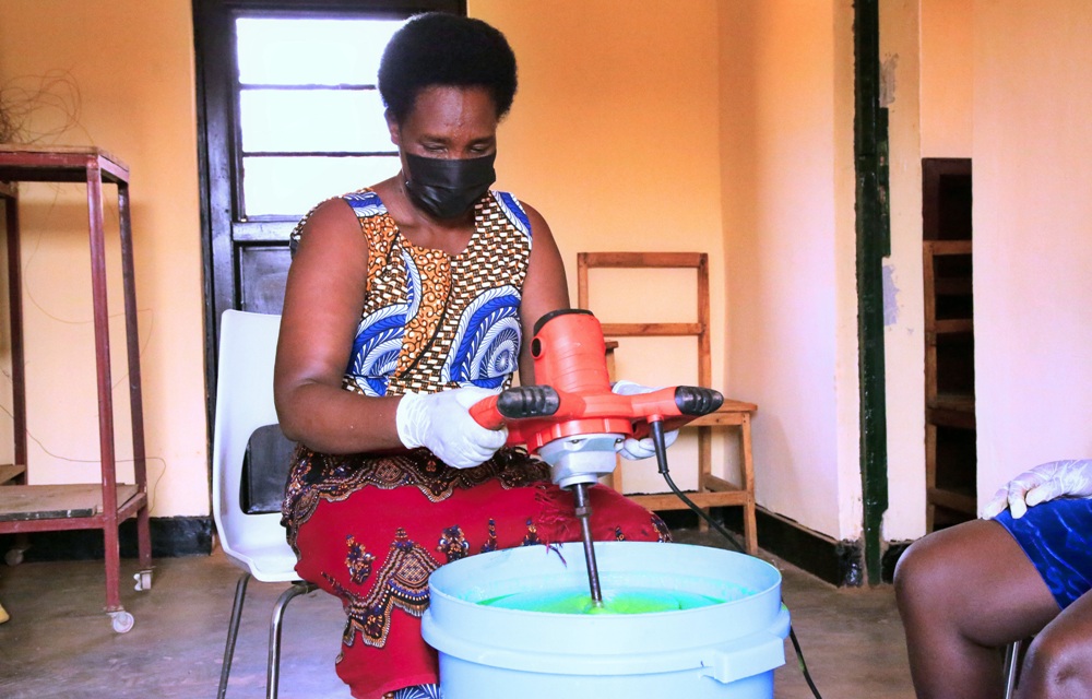 Woman wearing a mask and gloves uses a red handheld mixer to stir a bright green liquid in a blue bucket indoors.