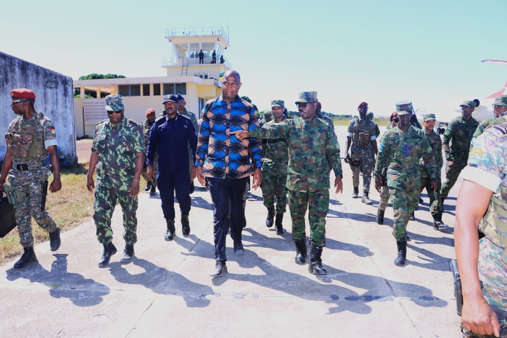 Man in a blue patterned shirt walking with camouflaged soldiers on a sunny airstrip, control tower in back.