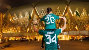 Teal Saudi Arabia soccer jersey being lifted on shoulders in front of a glowing gold-lit modern building at night, celebration scene.