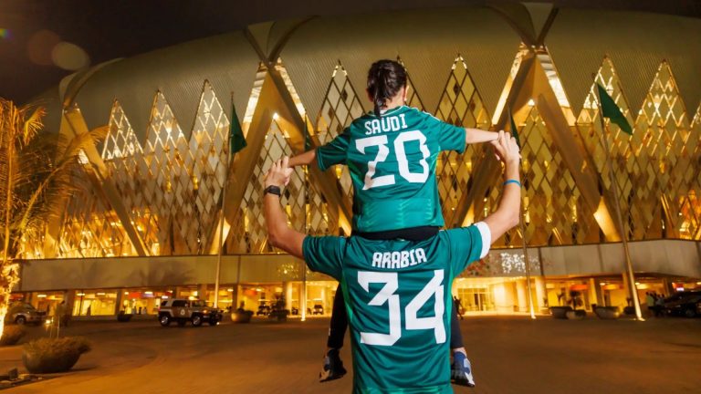 Teal Saudi Arabia soccer jersey being lifted on shoulders in front of a glowing gold-lit modern building at night, celebration scene.
