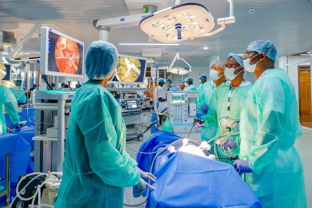 Medical team in green gowns and caps perform a live surgery under bright overhead lights, using monitors and anesthesia equipment in a modern operating room
