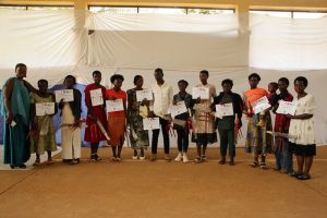 Group of children and two adults stand in a hall, each child holding a certificate during an award ceremony.
