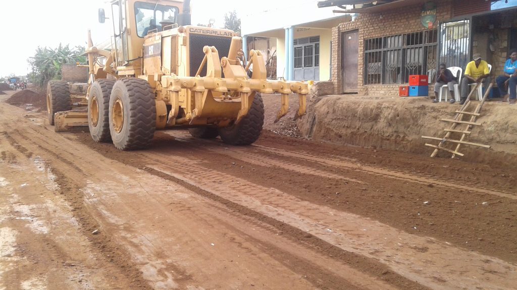 Large yellow Caterpillar road grader on a dirt street next to a row of houses; people sit outside watching the machinery.