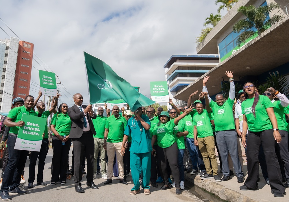 Group of people in green shirts marching outdoors, holding signs that read Save, Invest, Insure, with a man waving a large green flag.