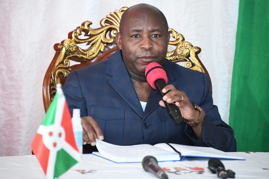 Man in a dark suit speaking into a pink microphone at a press conference, Burundi flag visible on the table in front of him