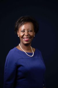 Smiling Black woman in a royal blue dress with a pearl necklace and earrings, against a dark studio background.