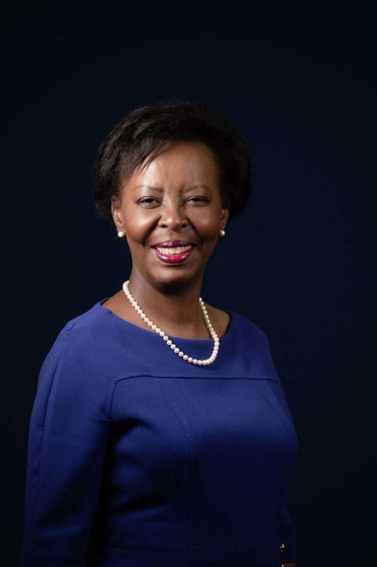 Smiling Black woman in a royal blue dress with a pearl necklace and earrings, against a dark studio background.