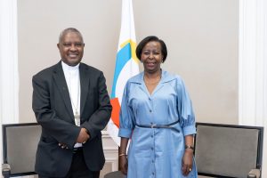 Two formally dressed individuals pose for a photo in a room with a white flag featuring blue and yellow shapes behind them, possibly a formal meeting setting.