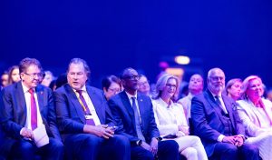 Group of professionally dressed attendees seated in a row at a conference under blue lighting.