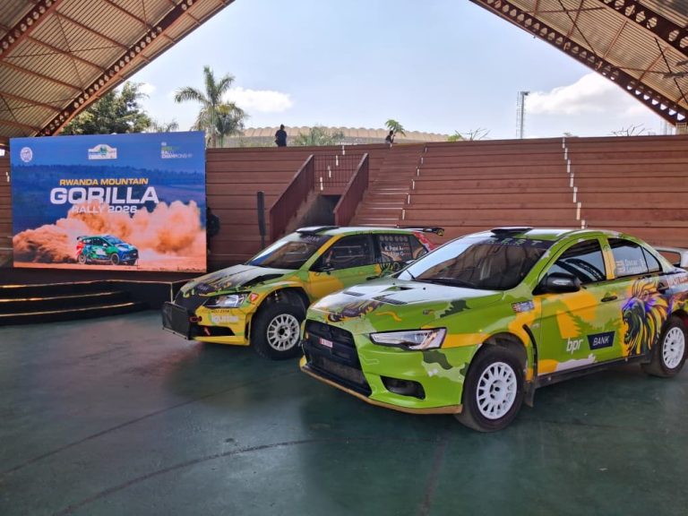 Two brightly colored rally cars are parked in a covered venue beside a large blue banner that reads 'Rwanda Mountain Gorilla Rally 2026'.