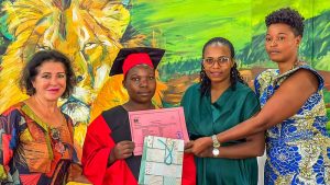 Graduate in a red cap and gown receives a pink diploma from three smiling women against a colorful mural backdrop.