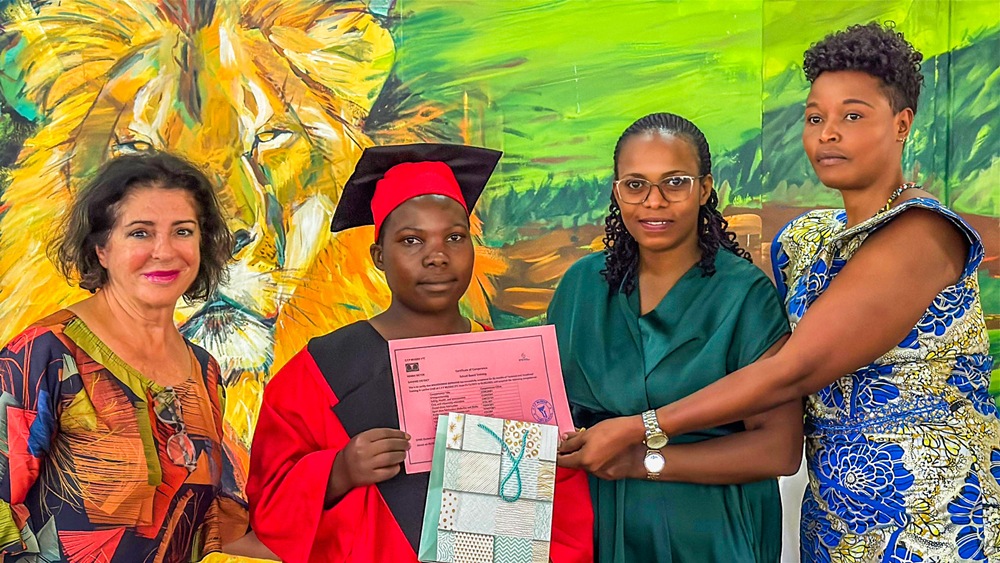 Graduate in a red cap and gown receives a pink diploma from three smiling women against a colorful mural backdrop.