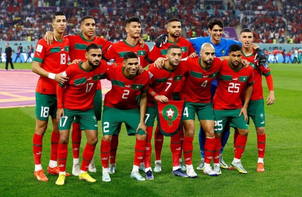 Morocco national football team posing for a group photo on the field in red shirts and green shorts with stadium crowd behind them.