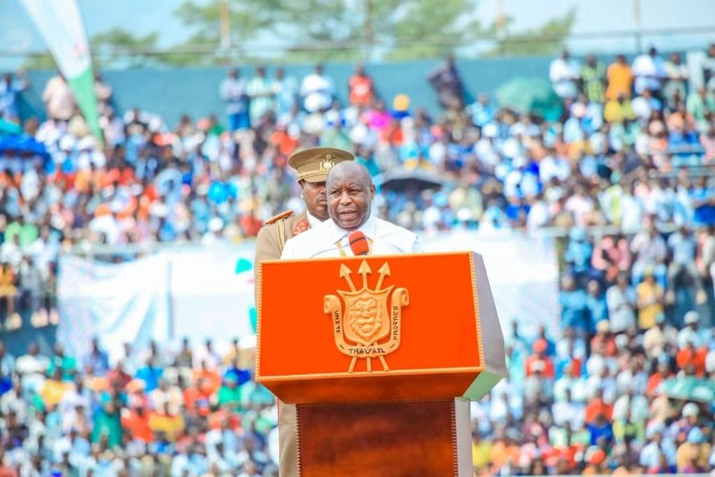 Public speaker at a podium giving a speech to a large stadium crowd with a crest on the front of the podium