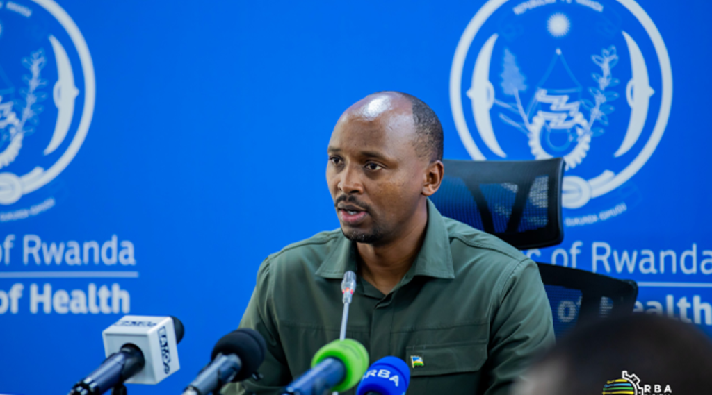 Man in a green shirt speaks into a microphone at a press briefing, with blue Ministry of Health backdrop and logos behind him.