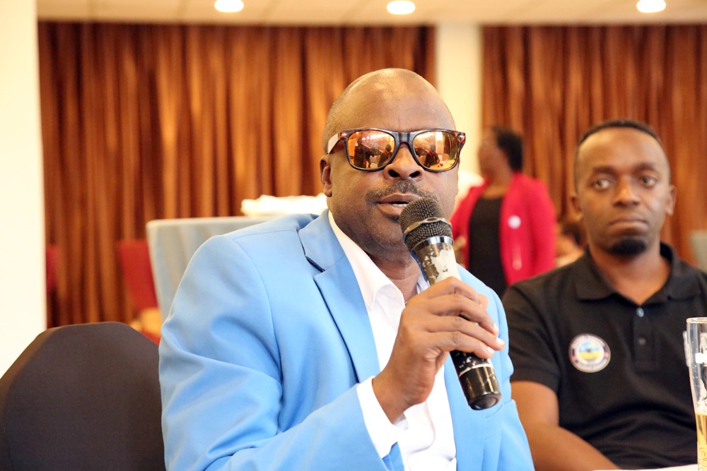 Man in a light blue suit and white shirt, wearing sunglasses, speaks into a handheld microphone at a formal event. A man in a black polo sits in the background.