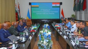 Delegates from India and Rwanda sit around a long conference table for a defense meeting, with national flags and a large screen in the background showing the meeting title.