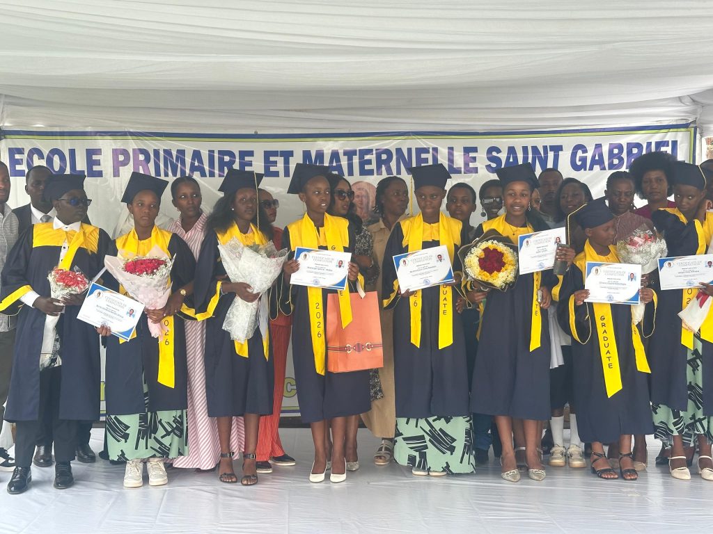 Group of graduates in blue gowns and yellow sashes, holding diplomas and flowers on stage under a school banner