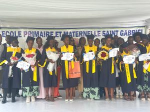 Group of graduates in blue gowns and yellow sashes, holding diplomas and flowers on stage under a school banner