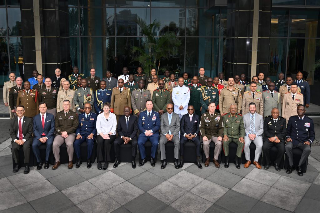 Group of military officers and officials posing for a formal photo outside a glass-front building on steps outside a corporate complex.