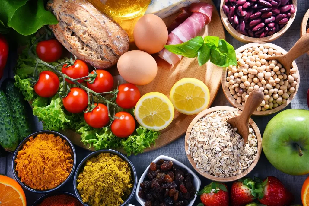 Colorful collage of fresh produce and pantry staples: tomatoes on the vine, greens, bread, eggs, lemons, apples, beans, oats, and spices on a wooden surface.